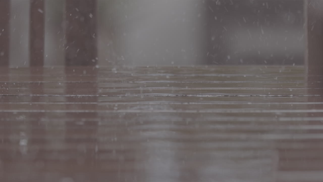 Close up shot of rain drops falling on a wooden table making splashes with chairs in the background in slowmotion LOG