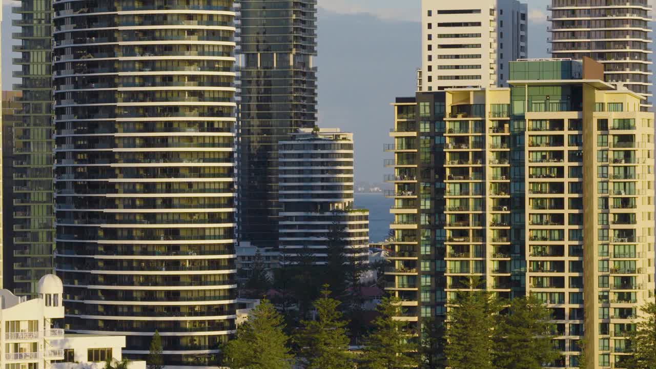 Drone footage captures Gold Coast skyscrapers with evening light casting shadows. The urban landscape blends with natural surroundings