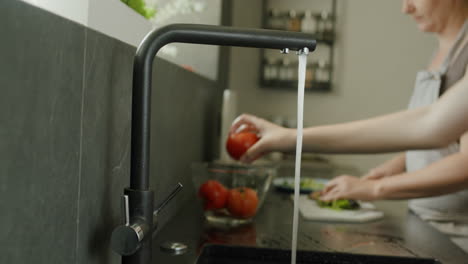 Mom-and-daughter-are-preparing-a-salad-in-the-kitchen.-In-the-foreground,-hands-are-washing-a-salad,-in-the-back,-a-woman-cuts-them-on-a-cutting-board