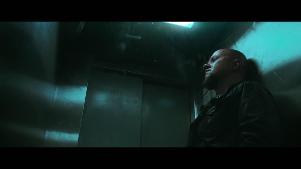 A bald dangerous looking man prepares a gun with the silencer in the elevator
