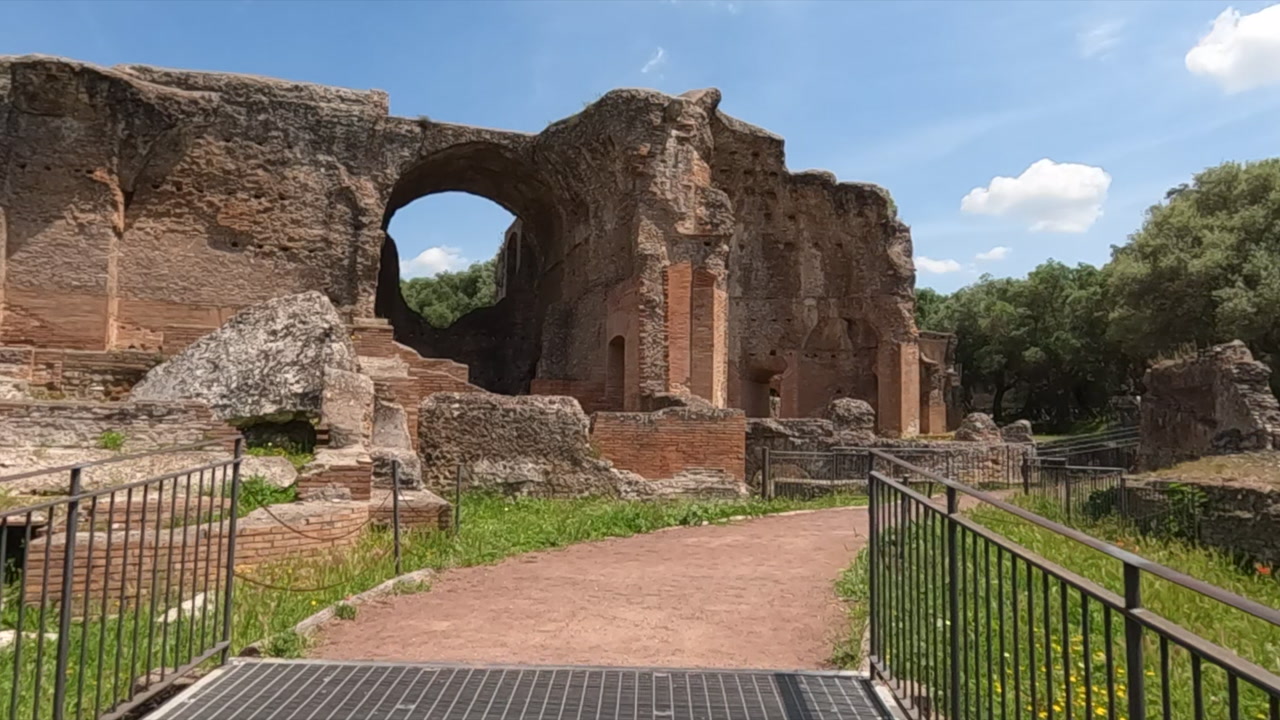 Panoramic view of crumbling ancient Roman ruins at Villa Adriana, Tivoli, near Rome. Massive brick arch, weathered stone walls, and lush greenery under blue sky.