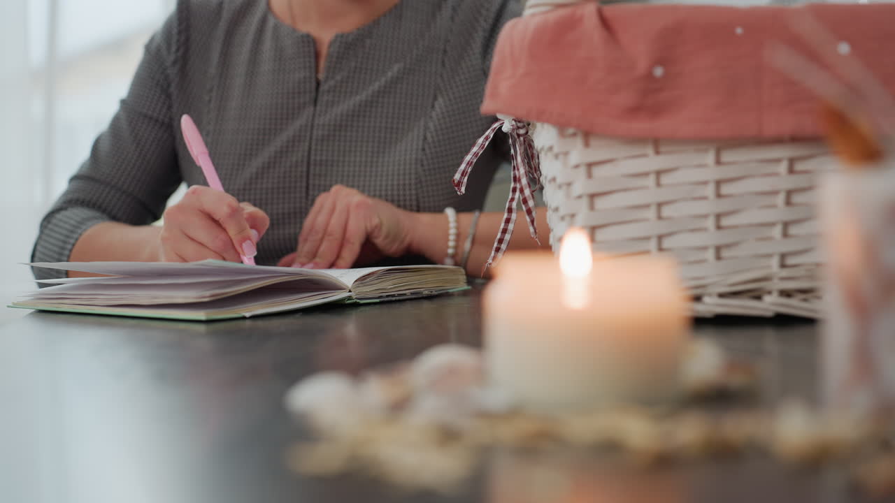 Blurred view of candlelight burning with seashells scattered on table while student sits writing in book using pink pen beside basket in calm cozy room filled with soft natural light