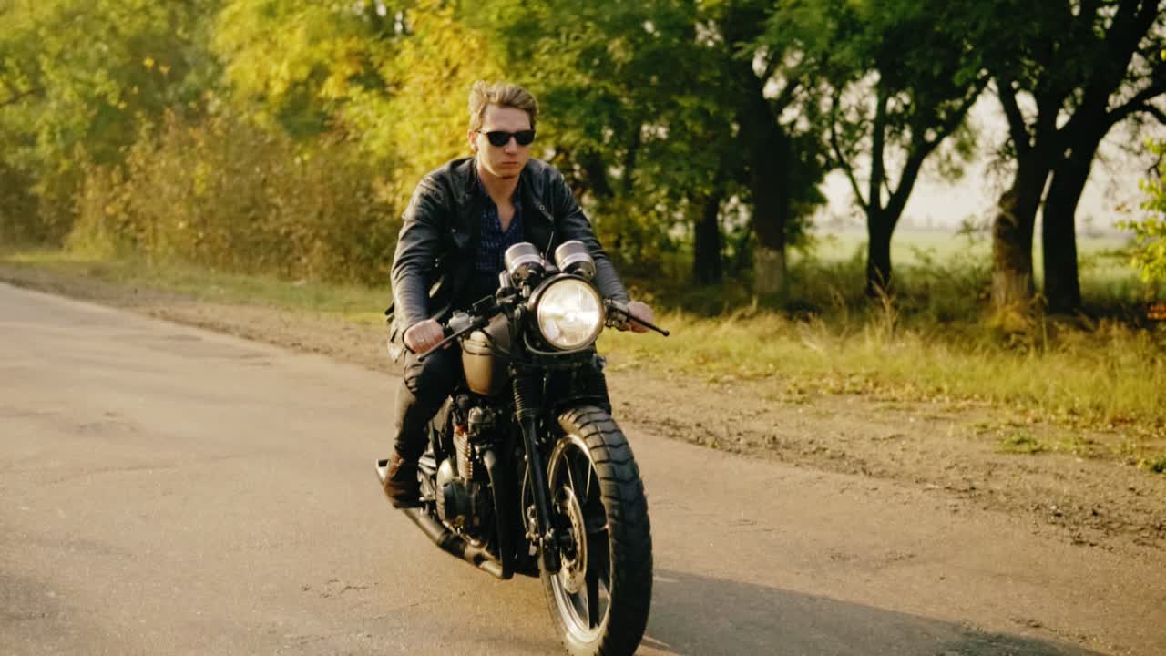 hombre elegante con gafas de sol y chaqueta de cuero montando su bicicleta fresca en y carretera de asfalto en un día soleado en el bosque en otoño