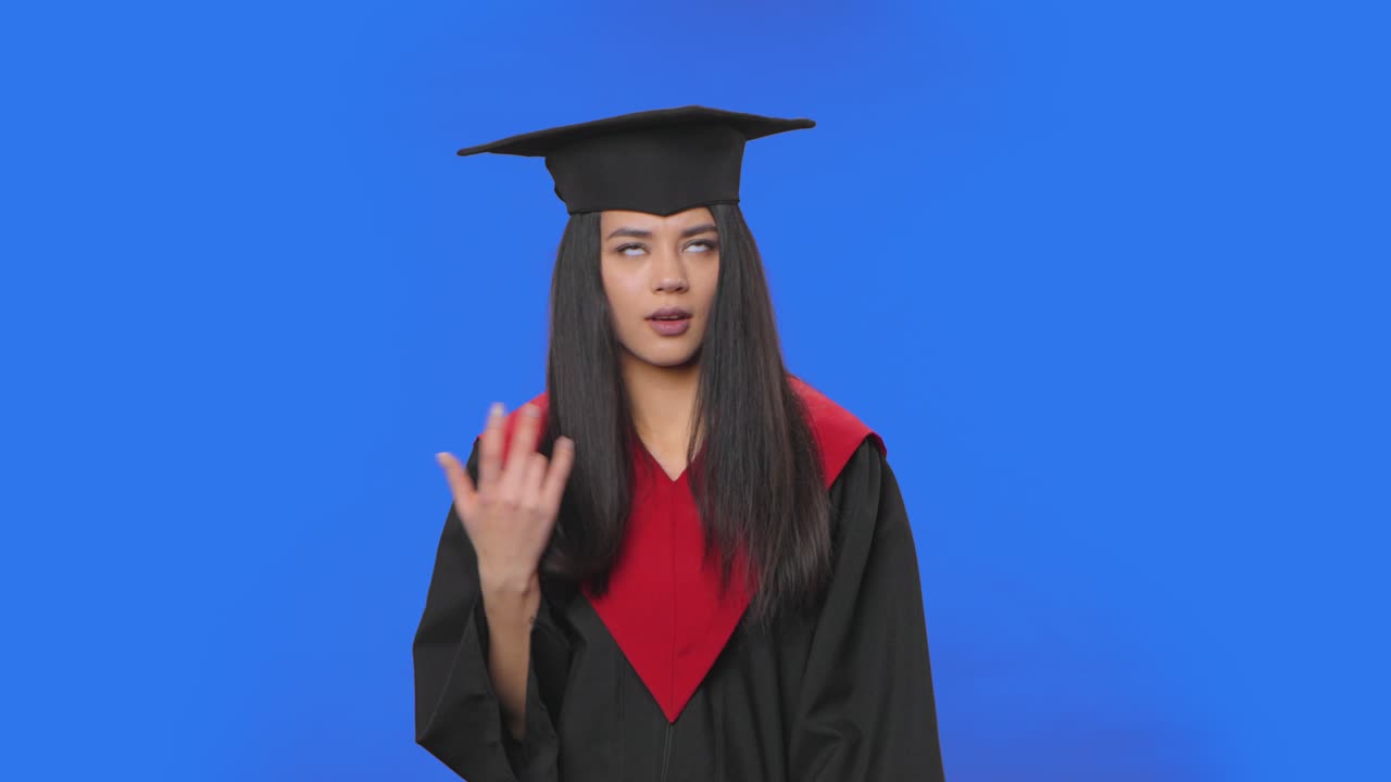 Portrait of female student in cap and gown graduation costume is indignantly talking to someone. Young brunette woman posing in studio with blue screen background. Close up. Slow motion ready 59.94fps