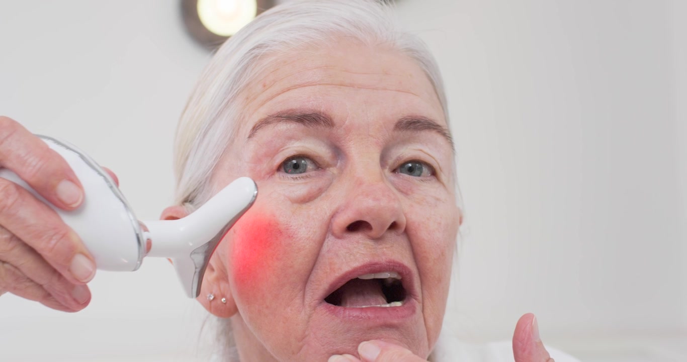Donna anziana che si tocca la pelle con un dispositivo LED rosso a tre punte che illumina, muovendolo sulle guance da sinistra a destra vicino al lavandino per la cura della pelle.