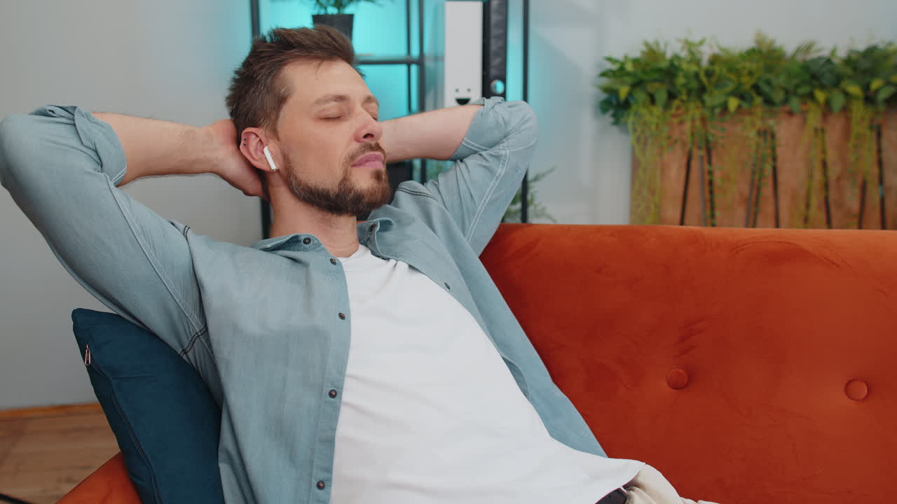 Giovane felice con le cuffie wireless che si rilassa sdraiato sul divano a casa ascoltando la sua musica preferita