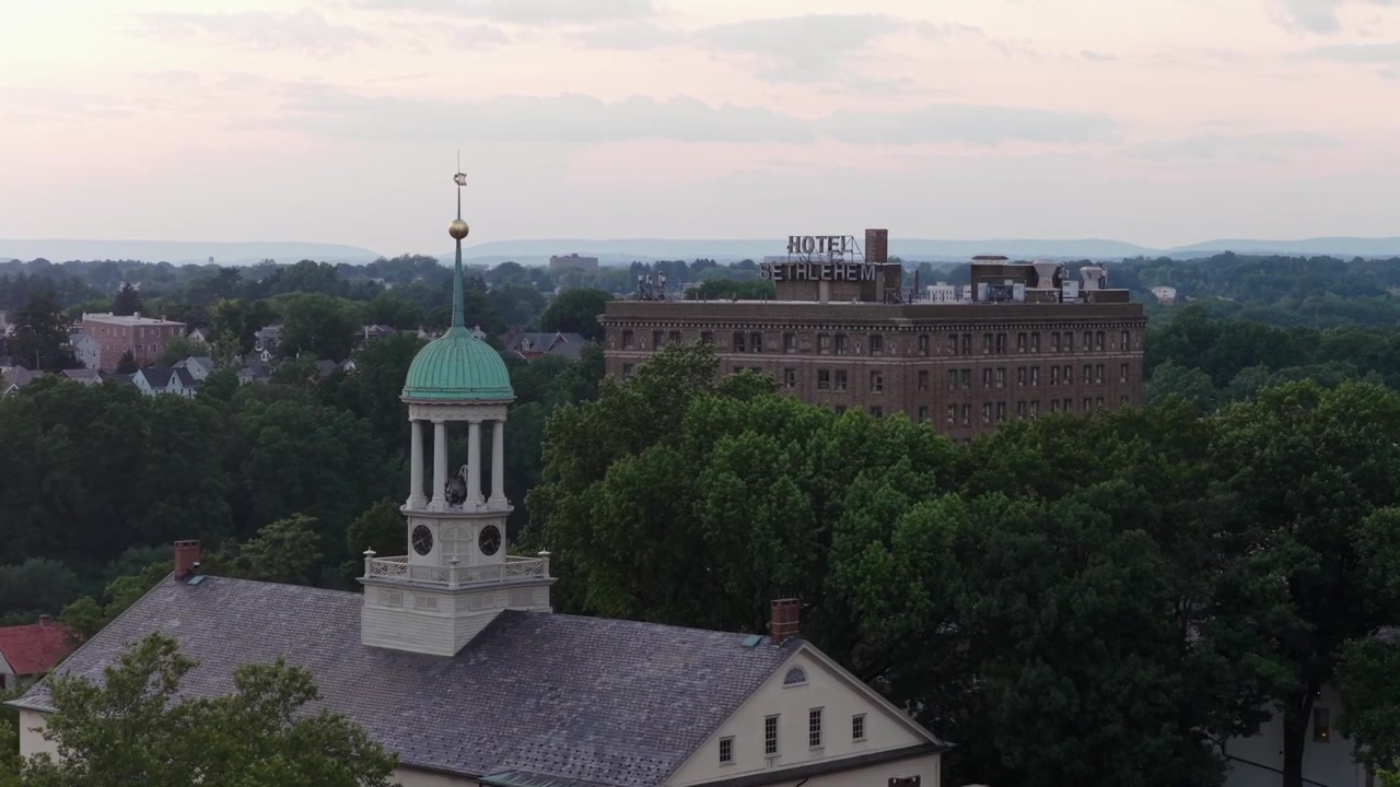 Campanile di chiesa bianco con cupola in rame verde e orologio si erge sopra alberi fitti vicino all'insegna dell'Hotel Bethlehem in cima a un edificio storico in mattoni al tramonto a Betlemme, Pennsylvania. Ripresa aerea con drone in avvicinamento