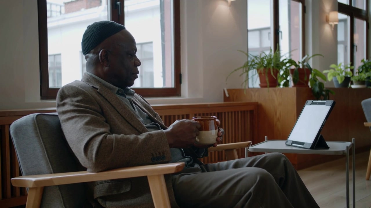 Noticing tablet, sipping from mug, man wearing cap, blazer leaning toward tablet in lounge watching