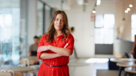 Portrait-Of-Smiling-Mature-Businesswoman-Standing-In-Busy-Open-Plan-Office