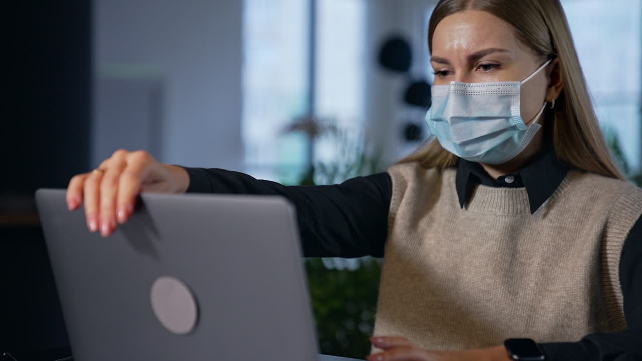 Female worker in protective mask looks disturbed at her laptop. Woman closes computer, takes off her mask and smiles into camera.