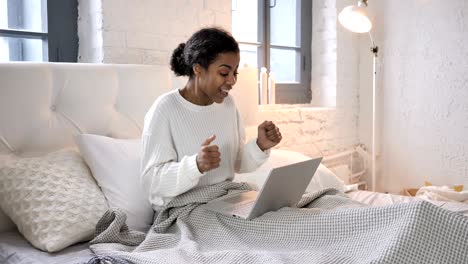 young african girl celebrating success on laptop while sitting in bed