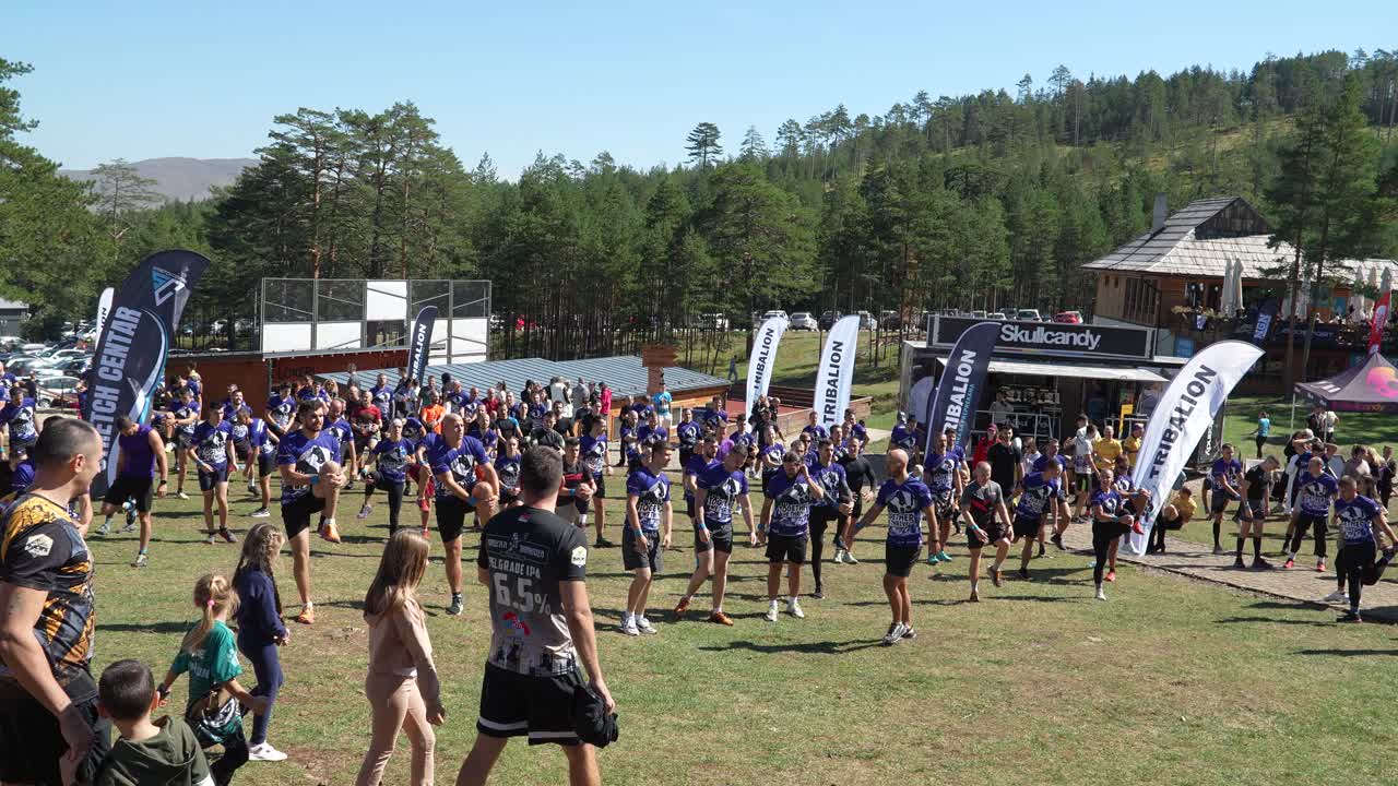 A large group of participants, wearing event shirts, are seen stretching and warming up on a grassy field surrounded by pine trees, preparing for a physically challenging outdoor event
