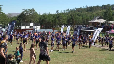 a large group of participants, wearing event shirts, are seen stretching and warming up on a grassy field surrounded by pine trees, preparing for a physically challenging outdoor event