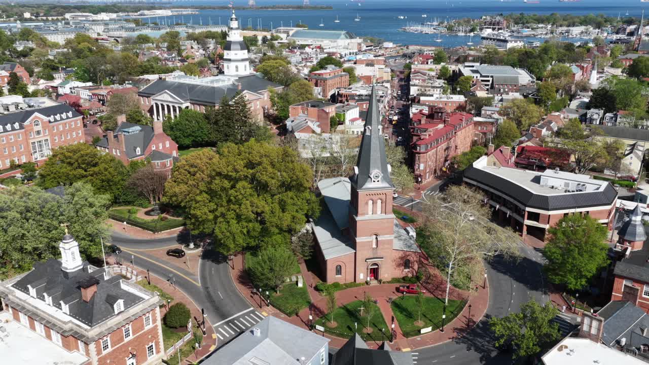 Órbita aérea de uma igreja histórica de tijolos no centro de Annapolis, Maryland, com ruas e árvores abaixo, enquanto a Casa do Estado de Maryland se eleva ao fundo sob o sol brilhante da primavera. Inclinação para cima.