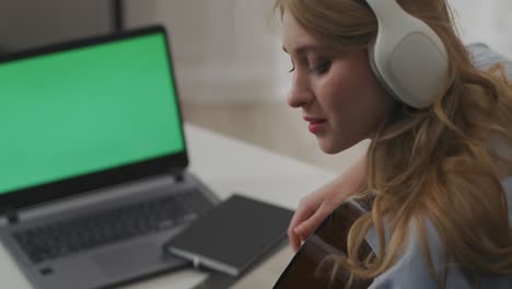 pretty female musician is playing guitar in front of web camera of laptop, online streaming concert from home, green screen