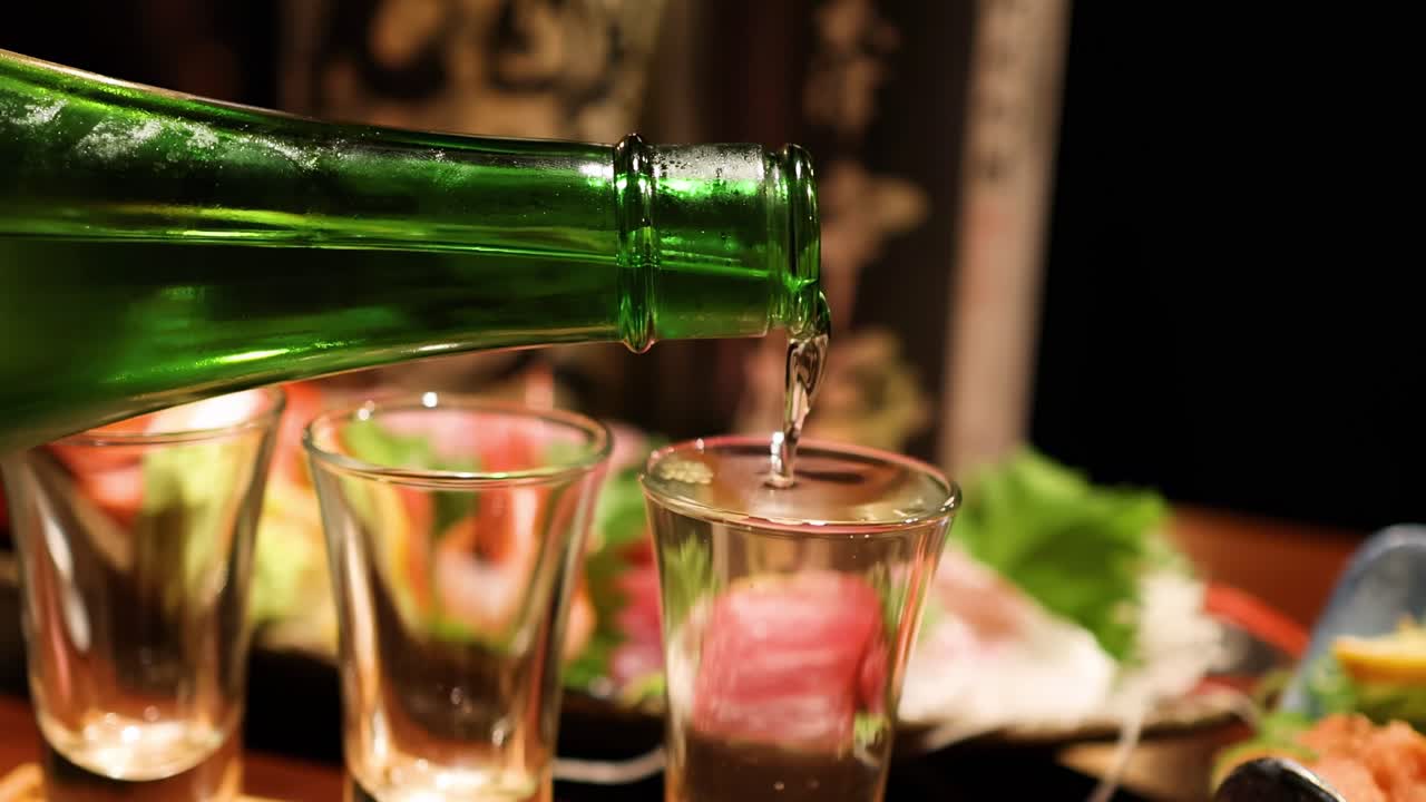 Close-up of sake being poured into glasses beside a sushi platter with vibrant garnishes.