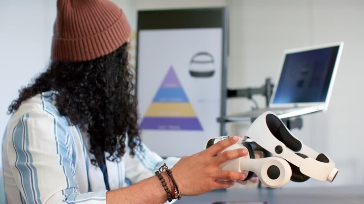 Hands entering from left, reaching for white VR headset on desk while designer checking quality