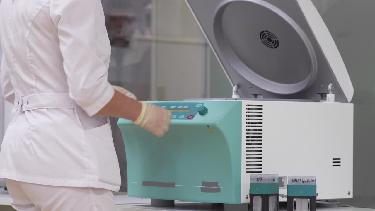 실험실에서, 목욕옷과 장갑을 입은 한 여성이 과도한 세포 층을 풀고 분리하기 위해 시험관에 인간 세포  ⁇ 플을 넣습니다.