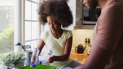 hija afroamericana jugando con botellas de plástico mientras clasifica el reciclaje con el padre en la cocina