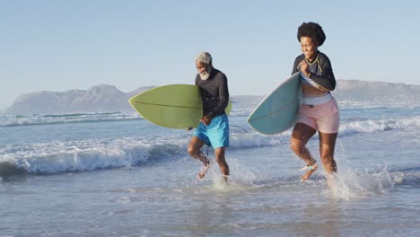 Glückliches-Afroamerikanisches-Paar,-Das-Mit-Surfbrettern-Am-Sonnigen-Strand-Läuft