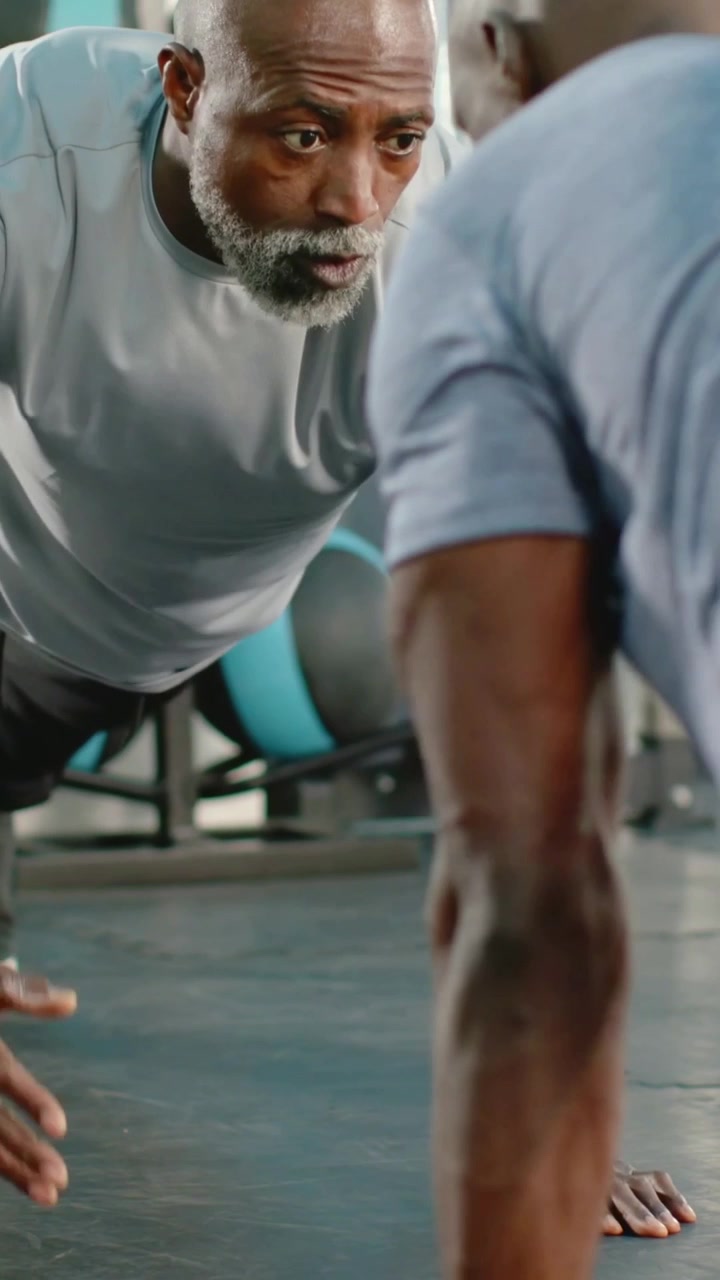 Vertical video: Cueing coach guiding trainee in blue shirt doing pushups at gym mat, teal med balls