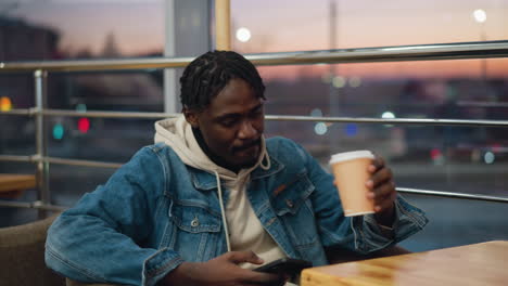 young man in denim jacket and beige hoodie relaxes in cozy cafe during sunset, holding smartphone in one hand and takeaway coffee cup in other, surrounded by warm lighting and tranquil atmosphere