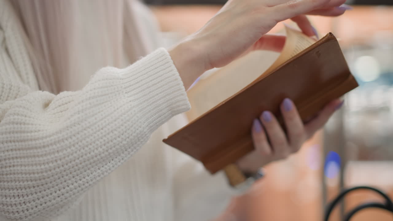manicured hands flipping pages of brown bound novel while wearing white knit sweater sleeve soft bokeh mall background warm light wristwatch visible black leather bag on bench