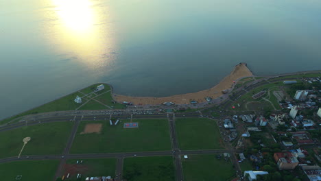drone tops down san josé beach at encarnación paraguay, sunset reflected on waterfront paraná river, green area and buildings