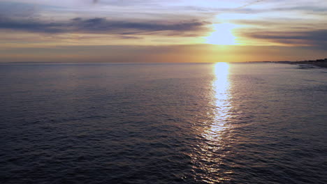 Cámara-De-Dron-Nivelada-A-Baja-Altitud-Retira-La-Toma-De-Carro-Con-Una-Puesta-De-Sol-Dorada-A-La-Vista:-Los-Surfistas-Debajo-Esperan-La-Ola-Perfecta-Y-Luego-El-Dron-Se-Eleva-Hacia-Arriba-Mientras-Vuela-Hacia-Atrás