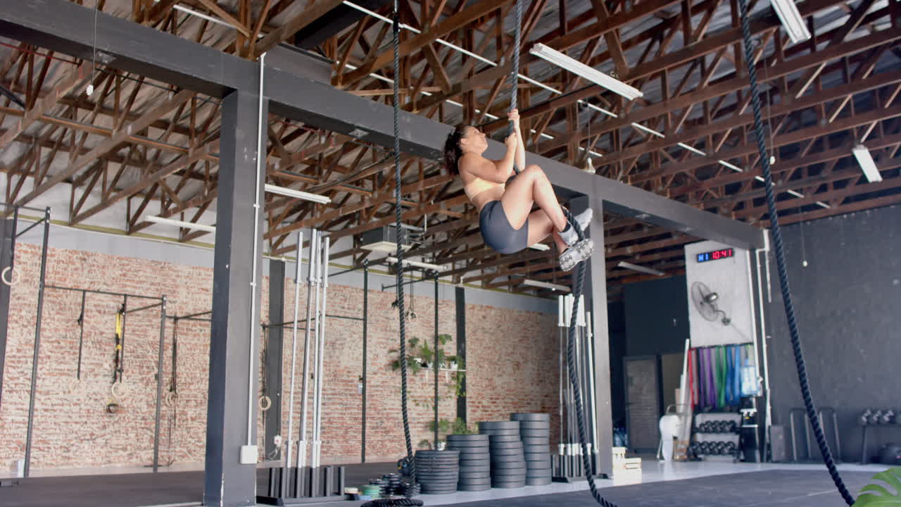 Performing rope climb exercise, strong fit woman training upper body strength in gym, copy space