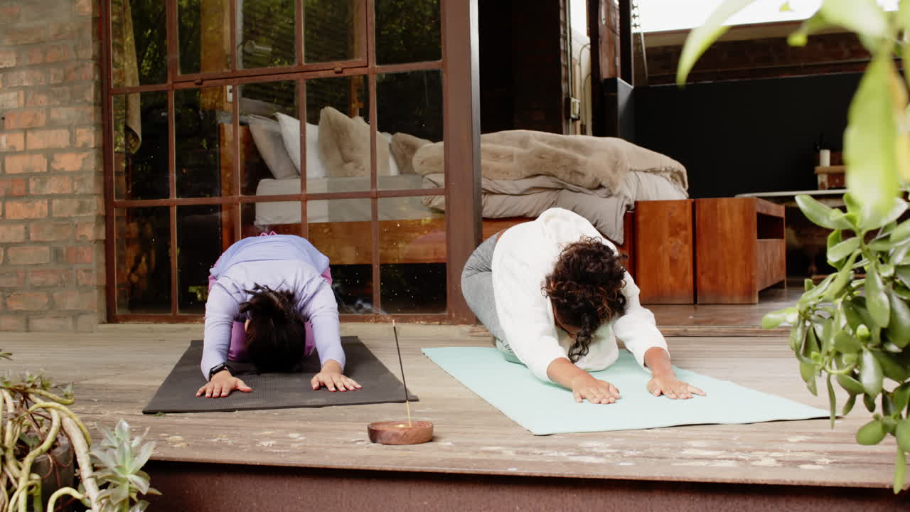 Multiracial female friends practicing yoga on porch, relaxing with incense and greenery around