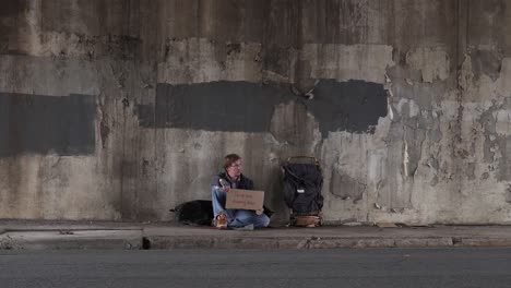 Wide-Shot-of-a-Homeless-Man-Sitting-on-the-Sidewalk-Holding-an-Anything-Helps-Cardboard-Sign