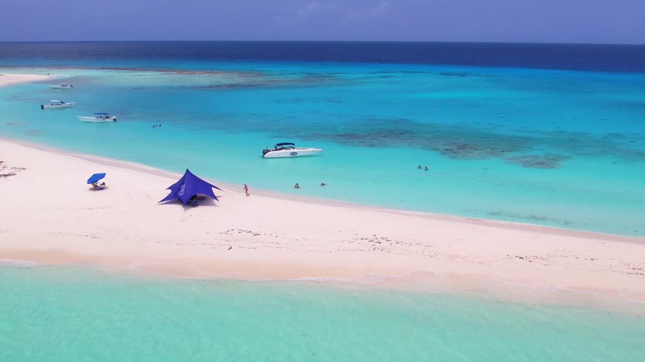 Zoom-out aerial of Cayo de Agua, Los Roques, Venezuela. Boats, people, and tent on white sand surrounded by turquoise Caribbean sea