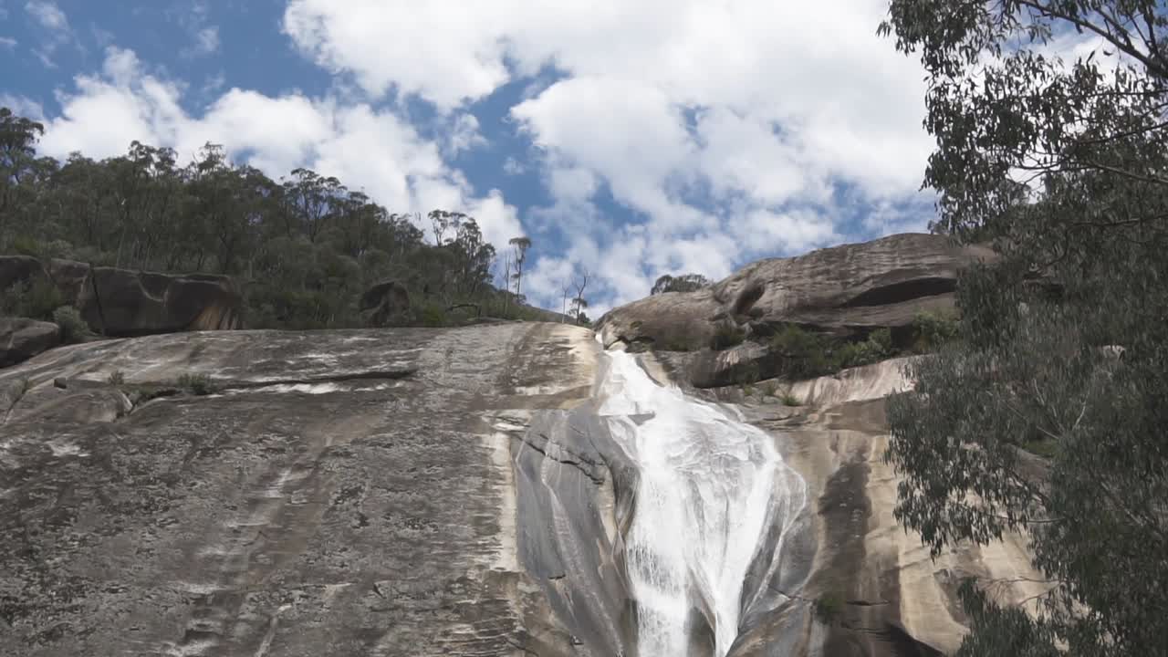 Stream water flows down from the forest across stones with beautiful clouds and blue sky