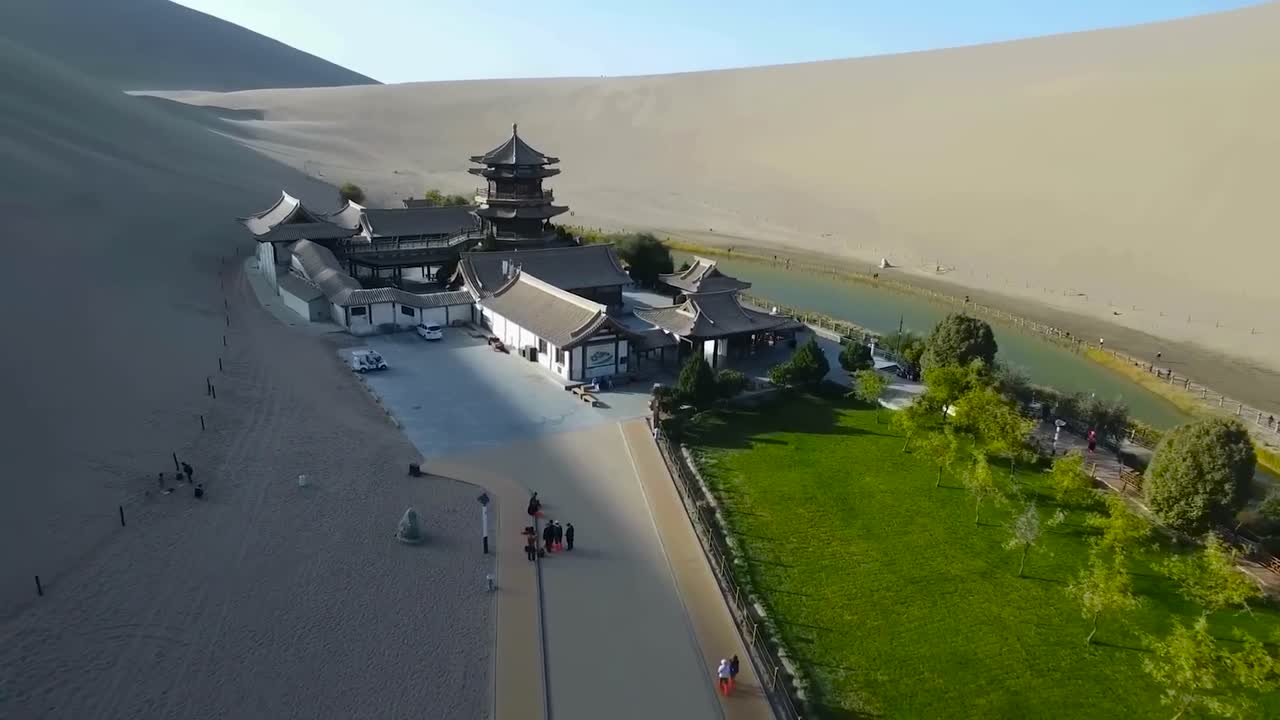 Aerial drone footage flying closer to Yueyaquan old architectural temple at Crescent lake in Chine during a sunny day. Tourist are seen walking below and sand dunes are surrounding the large building.