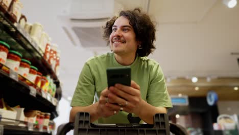 un chico moreno feliz con el cabello rizado mira videos divertidos en su teléfono mientras hace compras y conduce con un carrito en el supermercado