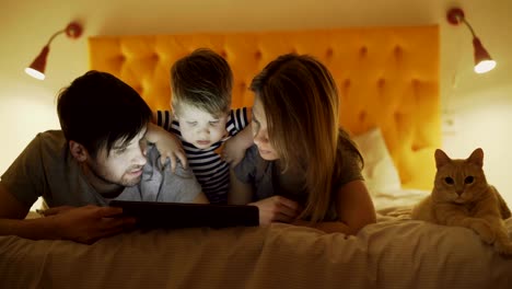 familia feliz con un hijo pequeño y un gato gracioso acostado en la cama en casa y usando una tableta para jugar antes de dormir