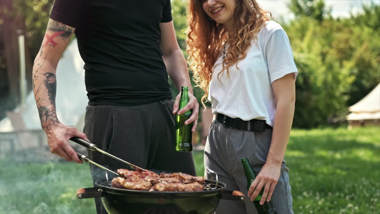 Giovane coppia che frigge carne alla griglia e beve birra. Tanto verde intorno. Glamping. Slow motion