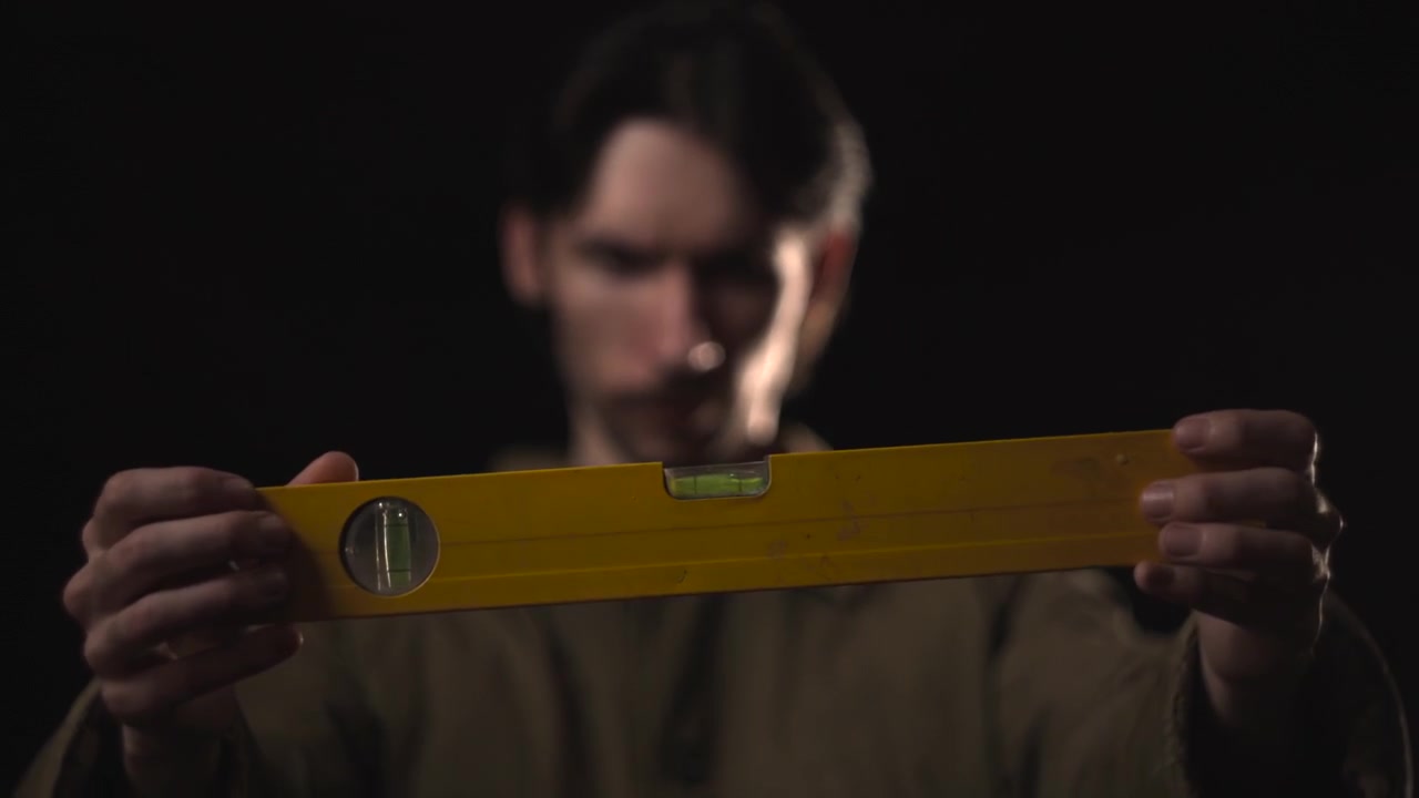 Un hombre difuminado se encuentra detrás de un nivel de burbuja amarillo sostenido horizontalmente en primer plano, con una iluminación de estudio oscura y poca profundidad de campo creando un fuerte