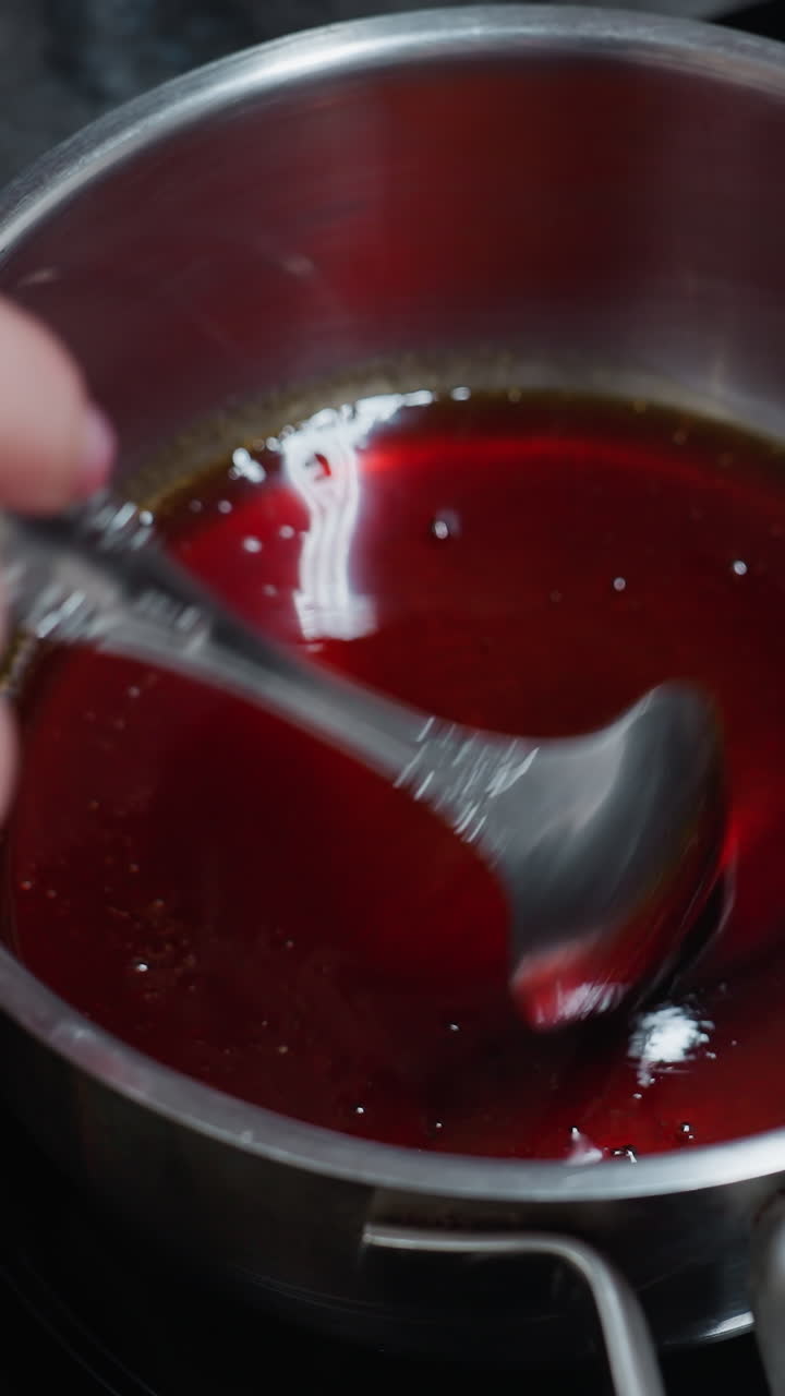 Close-up of white person hand stirring hot oil in stainless steel pot using a spoon on electric stove, with partial view of wooden tray and ingredients on kitchen countertop in background