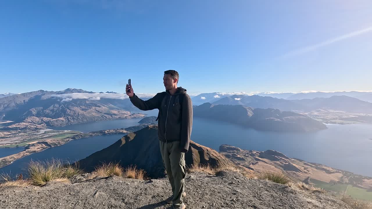 A man video calling from the top of Roy's Peak, a challenging hiking trail in Wanaka, New Zealand. This is an incredible view with beautiful mountains and lakes in the background
