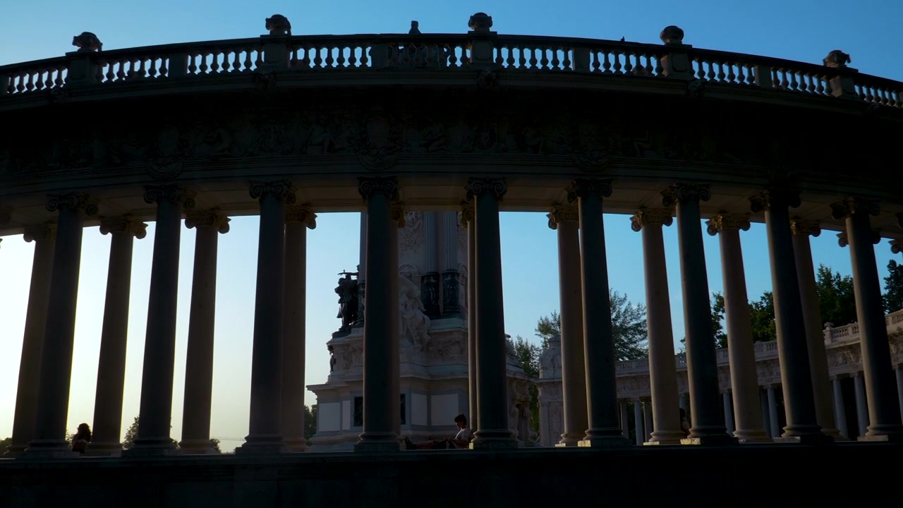 Una silhouette inquadrata dal basso dell'iconico colonnato semicircolare al Parco del Buen Retiro durante l'ora d'oro. Cattura l'architettura storica spagnola e le atmosfere di viaggio europee.