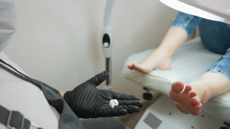 nail technician wearing black gloves pours cream into palm and applies it to client feet during pedicure session, with client resting on salon chair under lamp in bright clean environment