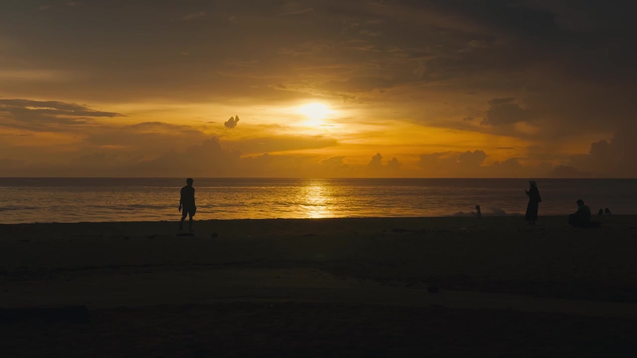 Slow motion silhouette scene of people relaxing on the beach during a dramatic orange cloudy sunset with warm evening light and peaceful ocean atmosphere.