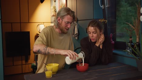 Un-Chico-Rubio-Con-Gafas-Y-Barba-Prepara-Un-Desayuno-Matutino-Con-Leche-Y-Cereales-Para-Su-Novia-Rubia-En-Una-Habitación-Acogedora