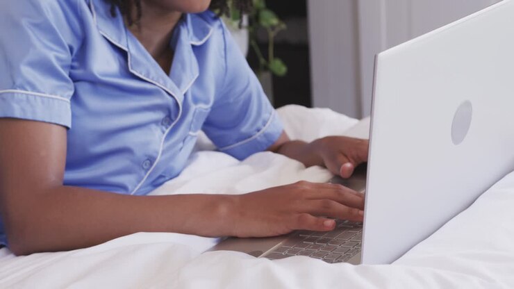Hands are typing on white laptop on bed, reading screen content and smiling while replying