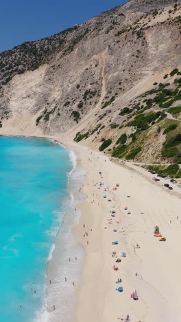 tourist sunbathing, swimming at myrtos beach surrounded by mountains and cliffs, kefalonia island, ionian sea, aerial descending shot, vertical