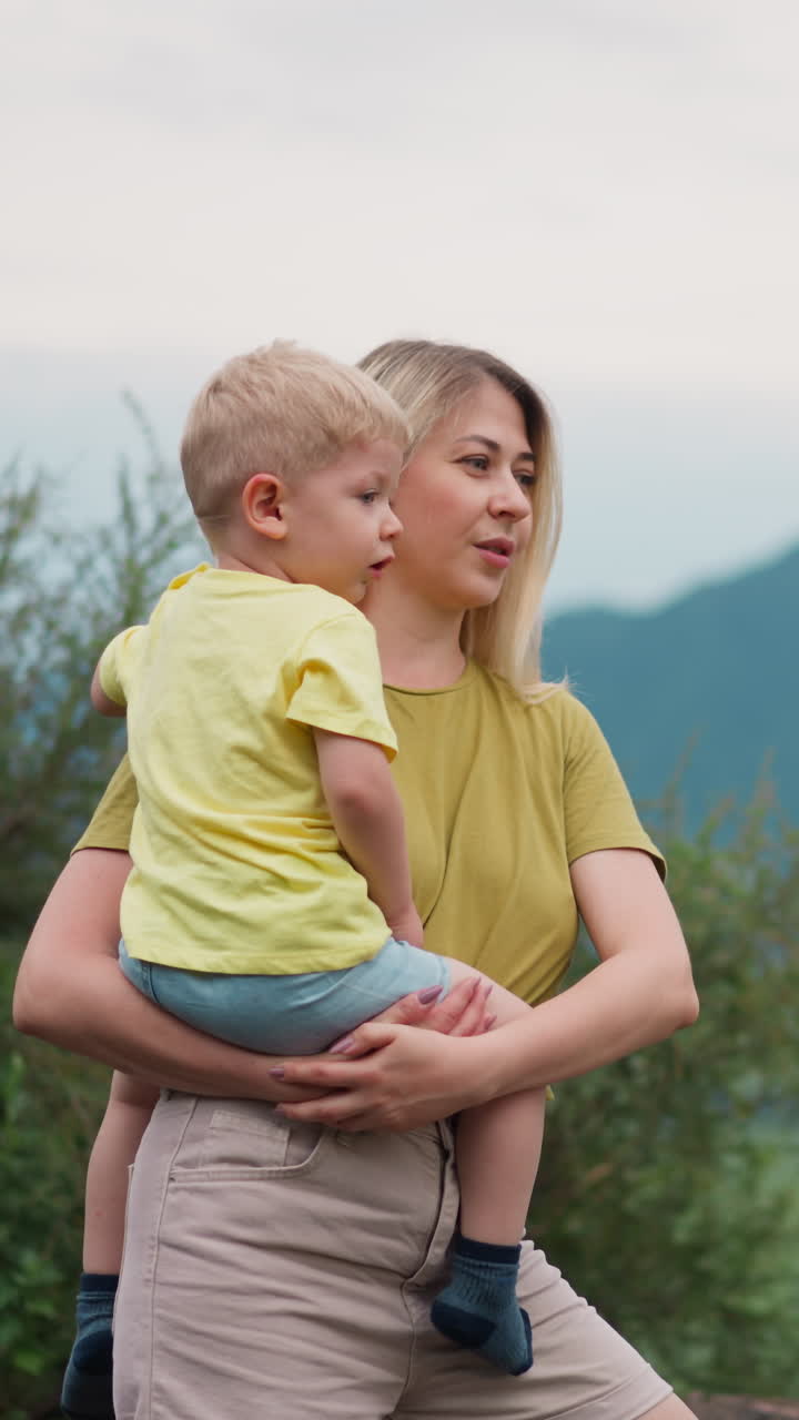 Happy woman with little son in arms enjoys picturesque view of river in valley canyon against mountains silhouettes at eco resort in summer slow motion