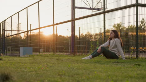 lonely woman near basketball stadium. young woman sits on grass in frustration suffering from loneliness at sunset. hard depressing time