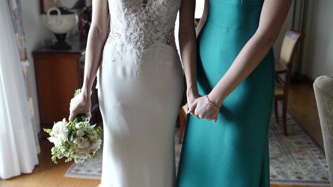 Bride in white dress holding bouquet stands beside woman in green dress holding hands inside room